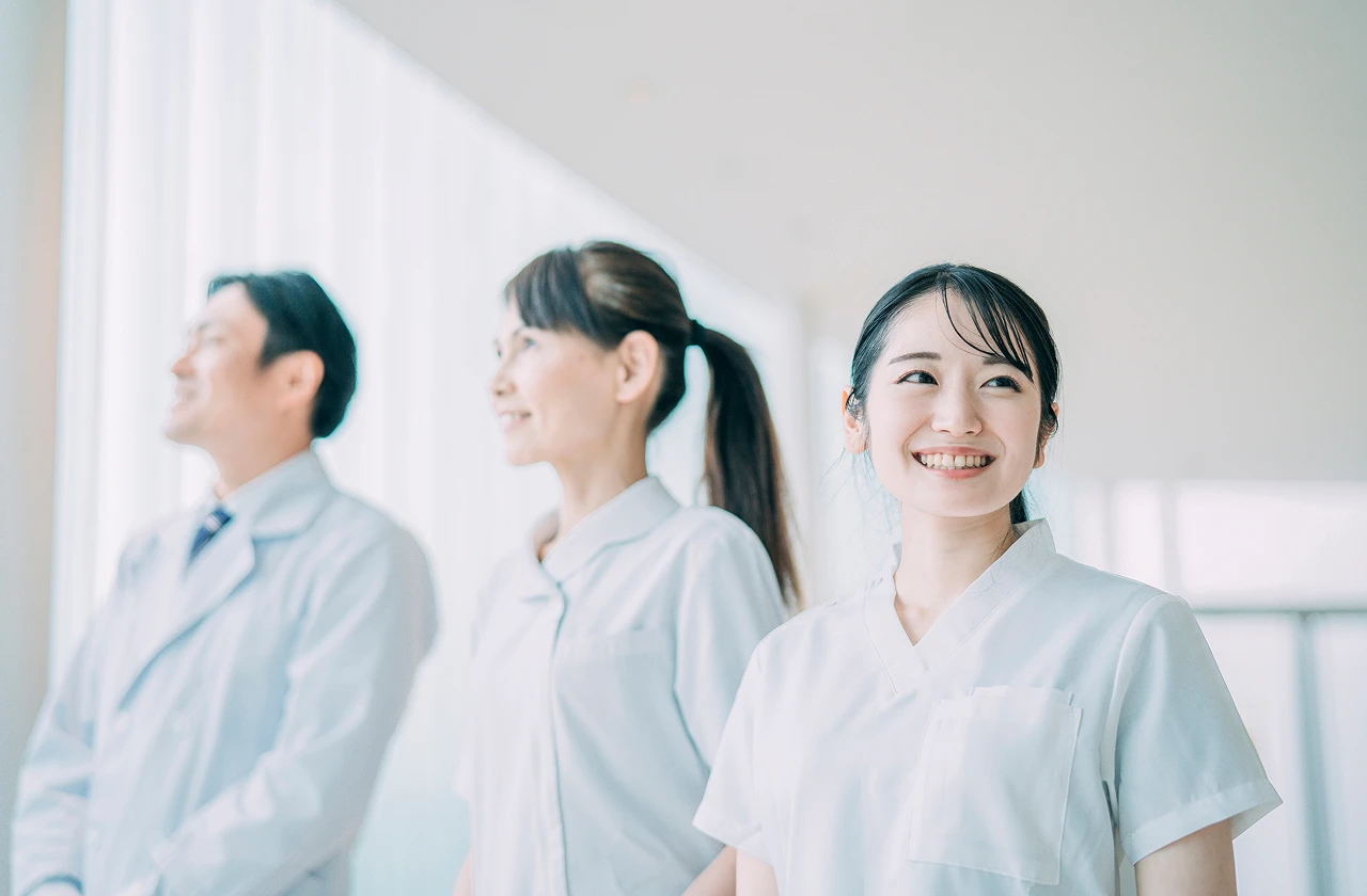 笑顔の⼥性スタッフを先頭に、医師や看護師が並んで明るい未来を⾒据える、活気とチームワークにあふれた職場のイメージ