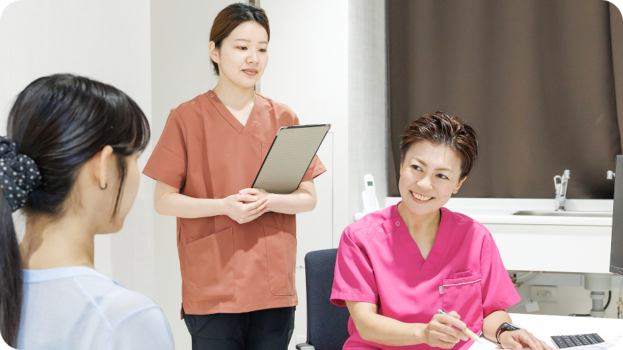 診察室で患者様のお悩みを笑顔で丁寧に聞き取る女性医師と、後ろで見守る医療スタッフの問診風景